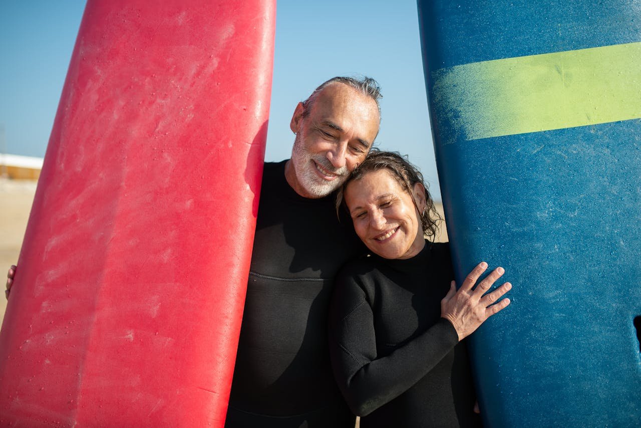 Smiling senior couple in wetsuits holding surfboards, enjoying a sunny day on a Portugal beach.